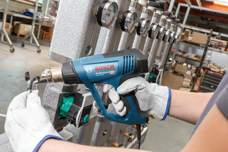 A person wearing safety equipment uses a heat gun on a cable near industrial gauges.
