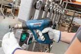 A person wearing safety equipment uses a heat gun on a cable near industrial gauges.