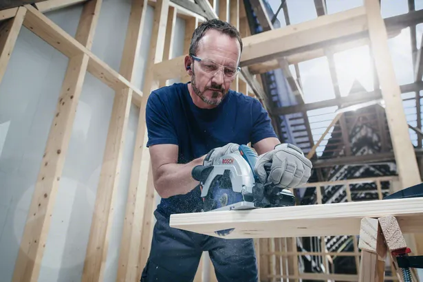 Person wearing safety equipment operates a circular saw to cut wood in a building under construction.