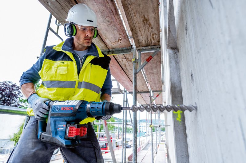 A person wearing safety equipment drills into concrete with a cordless rotary hammer.