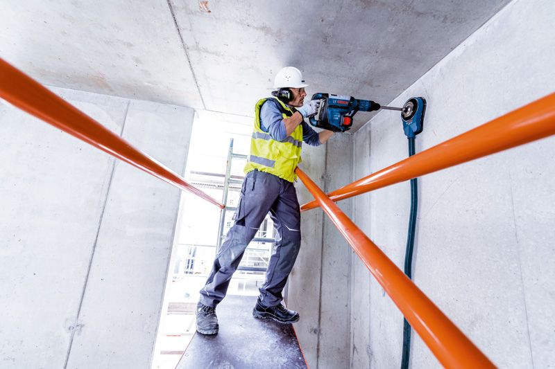 A person wearing safety equipment drills into a concrete ceiling with a rotary hammer.