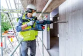 A person wearing safety equipment drills into concrete with a cordless rotary hammer.