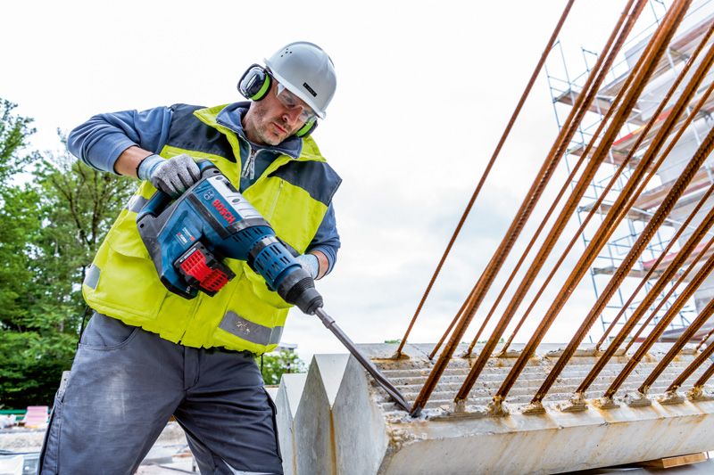 A person wearing safety equipment drills into concrete with a cordless rotary hammer.