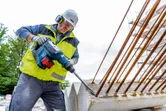 A person wearing safety equipment drills into concrete with a cordless rotary hammer.