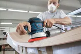 A person wearing safety equipment sands a curved wooden surface with a power tool.