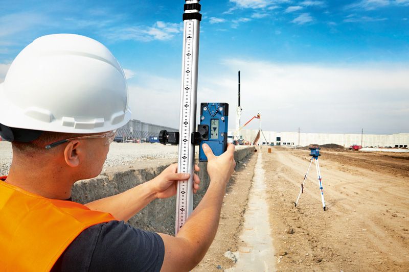 A person wearing safety equipment aligns a laser receiver with a laser leveling tool on a construction site.