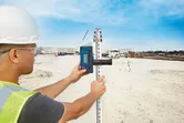 A person wearing safety equipment uses a laser receiver attached to a leveling rod at a construction site.