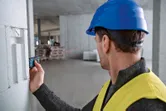 A person wearing safety equipment uses a laser measure against a concrete wall.
