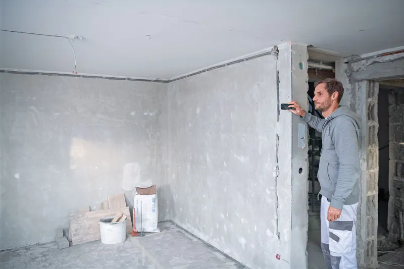 A person measures a wall in a construction site using a laser measure.