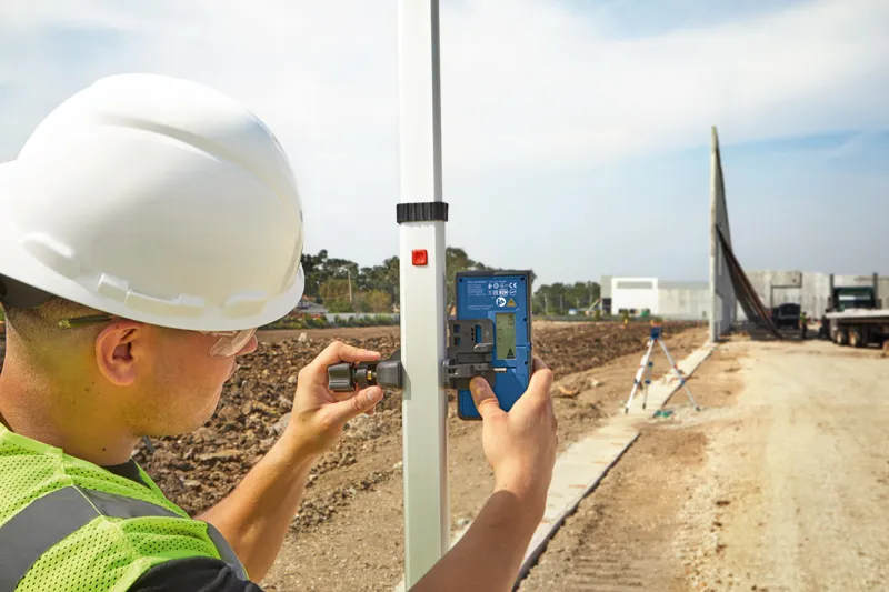 A person wearing safety equipment adjusts a laser receiver on a pole at a construction site.