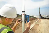 A person wearing safety equipment adjusts a laser receiver on a pole at a construction site.