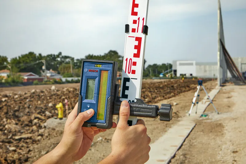 A person adjusts a laser receiver on a grade rod at a construction site.