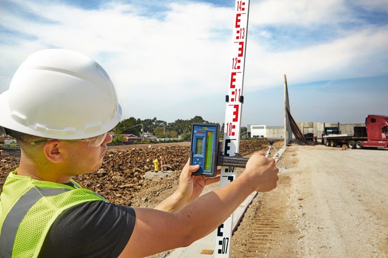 A person wearing safety equipment uses a laser receiver to measure a leveling rod at a construction site.