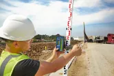 A person wearing safety equipment uses a laser receiver to measure a leveling rod at a construction site.