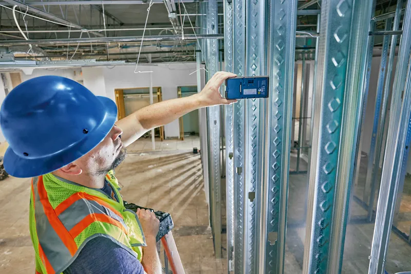 A person wearing safety equipment uses a laser receiver to check alignment on metal studs.