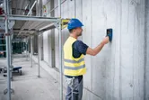 A person wearing safety equipment scans a concrete wall using a detector device.