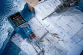 Electronic detector, drill, and fasteners placed on architectural blueprints.