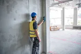 A worker wearing safety equipment uses a detector on a concrete wall.