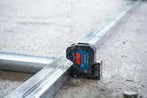 Laser leveling tool projecting green lines for aligning metal tracks on a concrete floor.