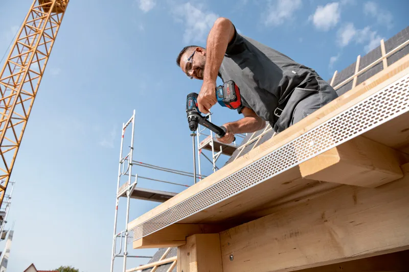 A person wearing safety equipment drills into a wooden beam on a building under construction.