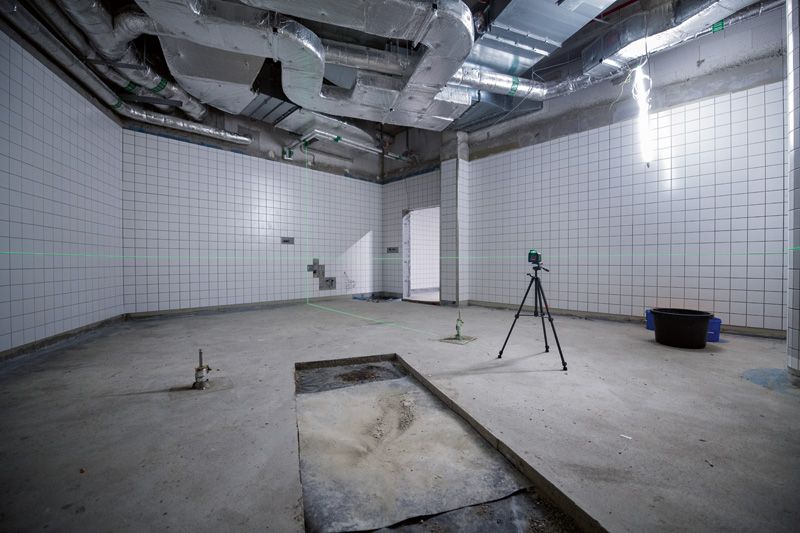 Laser leveling tool on a tripod projects green lines in a tiled room under construction.