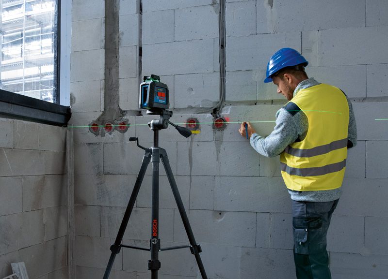A person wearing safety equipment marks a wall using a laser leveling tool on a tripod.