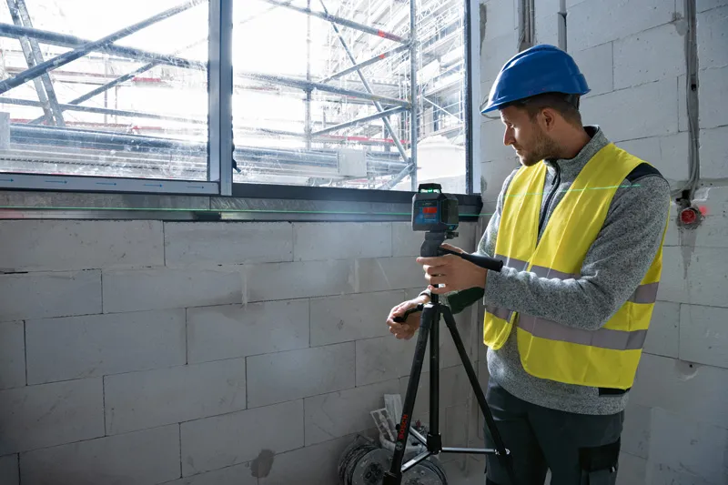 Person wearing safety equipment adjusts a laser leveling tool on a tripod near a window.