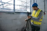 Person wearing safety equipment adjusts a laser leveling tool on a tripod near a window.