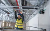 A person wearing safety equipment adjusts ceiling frames guided by a laser leveling tool.