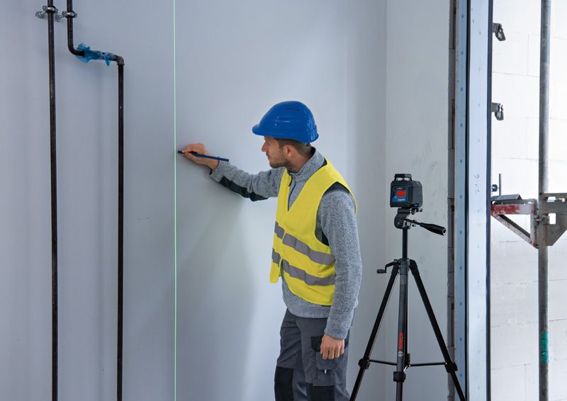 A person wearing safety equipment marks a wall using a laser leveling tool on a tripod.