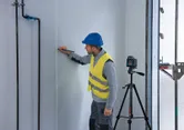 A person wearing safety equipment marks a wall using a laser leveling tool on a tripod.