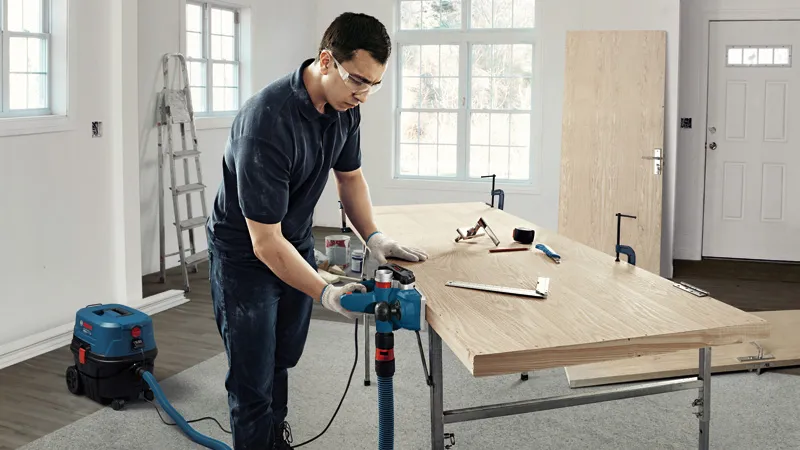 A person wearing safety equipment operates a planer on a wooden board in a workshop.