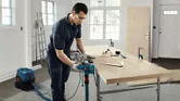 A person wearing safety equipment operates a planer on a wooden board in a workshop.