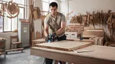A person wearing safety equipment planes a wooden board in a workshop.