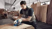 A person wearing safety equipment smooths a wooden plank with a planer in a workshop.