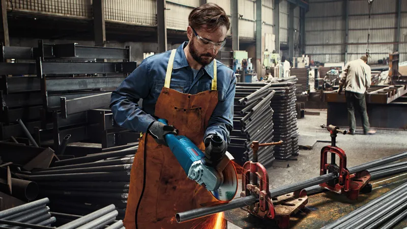 A person wearing safety equipment cuts metal pipe with a power tool in an industrial workshop.