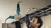 A person wearing safety equipment fastens drywall to a ceiling frame with a screwdriver.