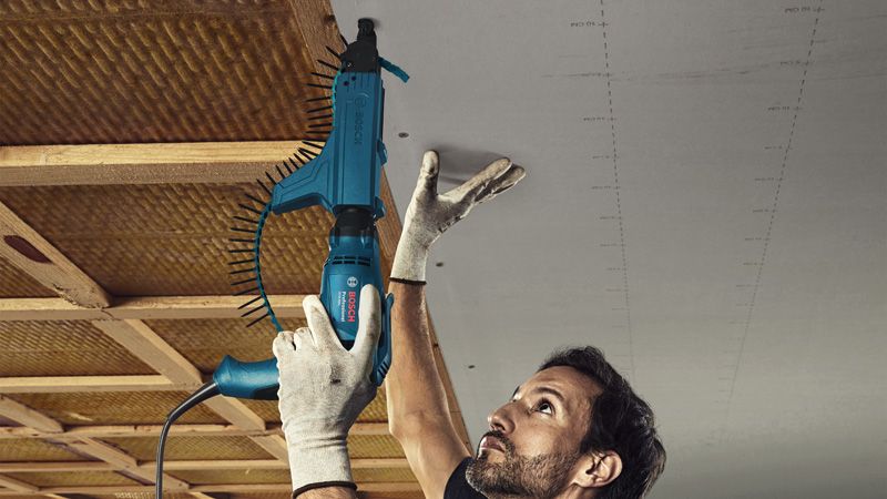 A person wearing safety equipment uses a drywall screwdriver to fasten a ceiling panel.