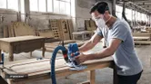 A person wearing safety equipment shapes a wooden panel using a router in a workshop.