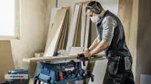 A person wearing safety equipment cuts wood on a table saw in a workshop.