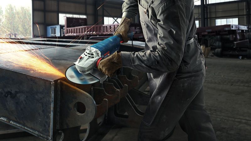 A person wearing safety equipment grinds a large metal beam with an angle grinder.
