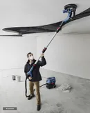 A person wearing safety equipment sands a ceiling with a drywall sander in a renovation space.