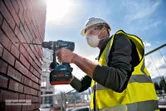 A person wearing safety equipment drills into a brick wall with a cordless combi tool.