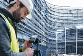 A person wearing safety equipment uses a laser measure on a tripod at a construction site.