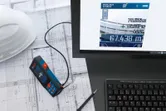 Laser measure connected to a computer on architectural plans beside a hard hat.