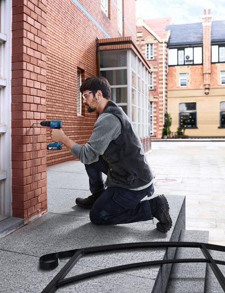 A person wearing safety equipment drills into a brick wall on a building exterior.