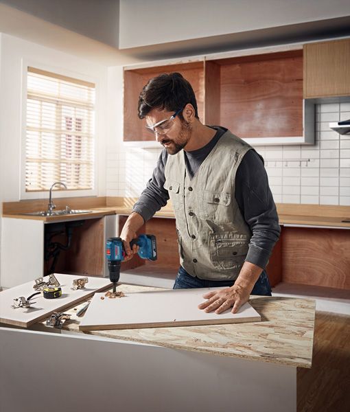 Person wearing safety equipment drills into a wooden board in a kitchen workspace.