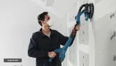 A person sands drywall with a power drywall sander on an unfinished wall.