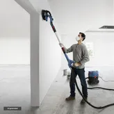 A person sands a white wall with a drywall sander connected to a dust extractor.