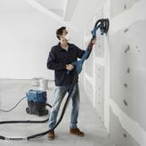 A person sands drywall with a drywall sander connected to a dust extractor.